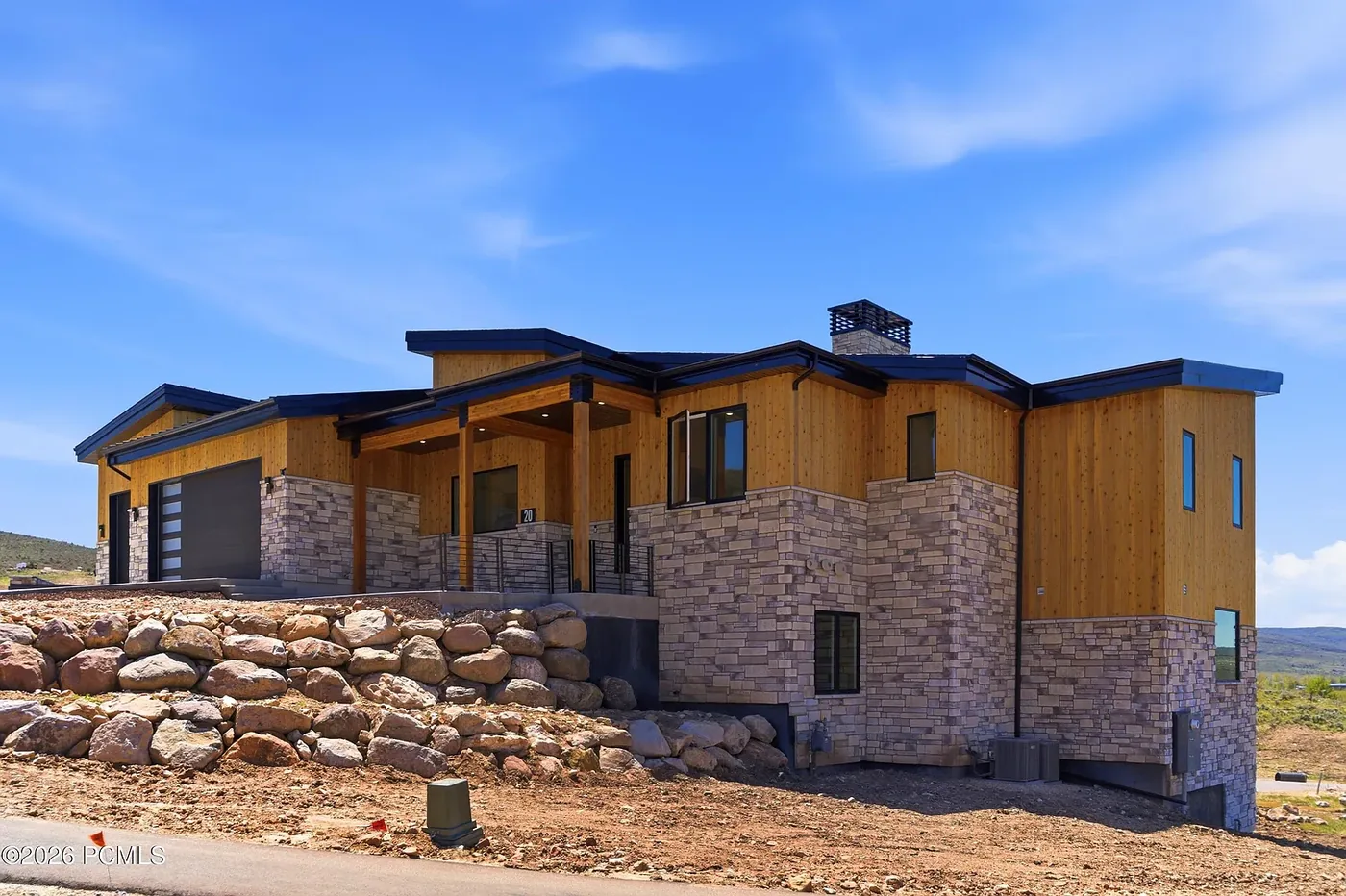 Modern mountain residence with cedar siding, stone base, and boulder retaining wall under blue sky